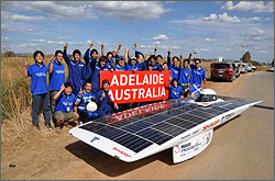 Photo of a team of young adults with their hands in the air and smiling, holding a sign that says 'Adelaide Australia.' In front of them is a solar car, a flat, low, square vehicle with a bubble near the rear for the pilot's head. The top of the vehicle is covered in solar cells.